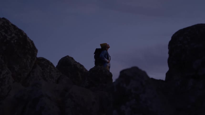 Hiker wearing a backpack stands on a mountain of rocks as she looks around