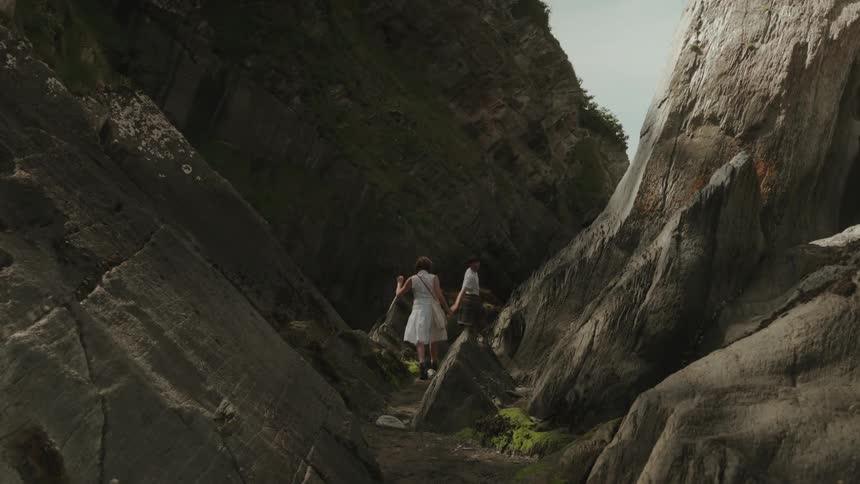 A young couple is climbing down a rocky path with the woman wearing a white dress and carrying a bag and the man wearing a white tshirt and shorts