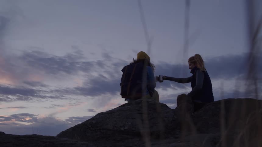 Two hikers are sitting on a rocky outcrop at dusk one wearing a yellow beanie and a backpack while the other hands her a cup as they talk under a cloudy purple and blue sky