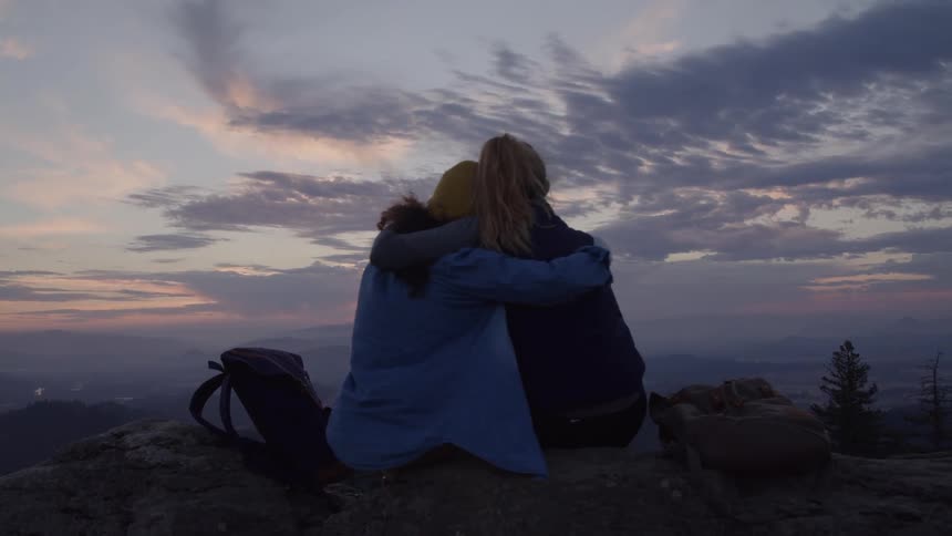 Two women sit on top of a hill watching the sun fall on the horizon then they hug and gently rock each other side to side