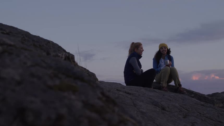 Two female hikers are sitting on a rocky mountain ridge at dusk with one wearing a yellow beanie and the other a dark blue vest as they talk and gesture to each other against a hazy cloudy sky