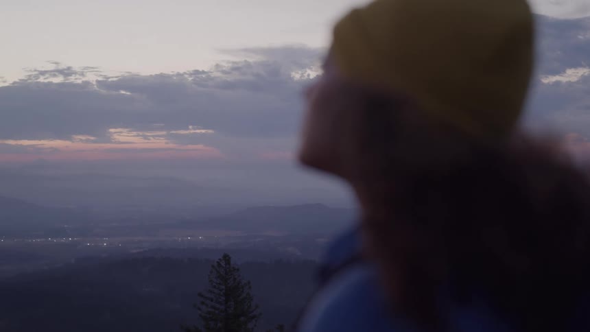 A woman wearing a yellow beanie is seen in a softfocus profile with a panoramic view of a valley with a town in the distance at dusk or dawn