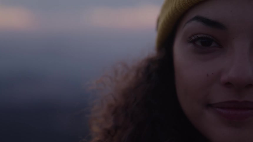 A closeup of a womans face shows the left half of her head with curly hair and a yellow beanie as she stand outdoors against a blurred background