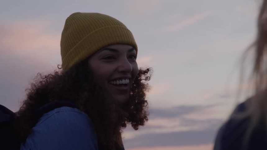 A young woman wearing a yellow hat with curly hair talks to another woman smiling at dawn