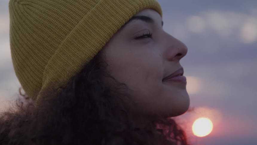 Woman wearing a yellow beanie smiles slightly while looking up during sunset