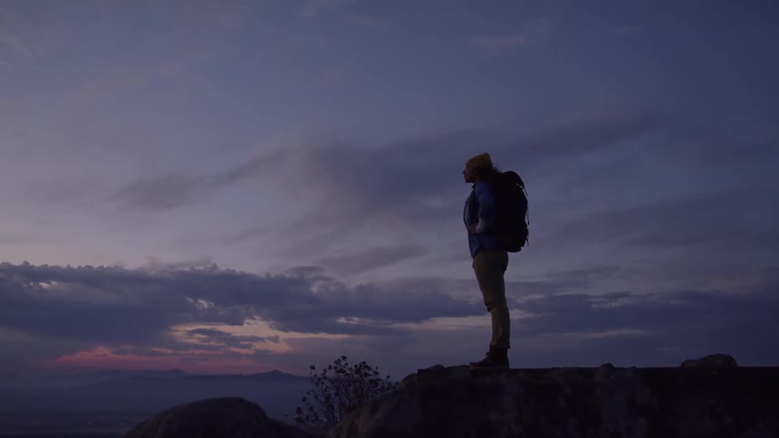 Person wearing a beanie and backpack standing on a rock watching the sunset
