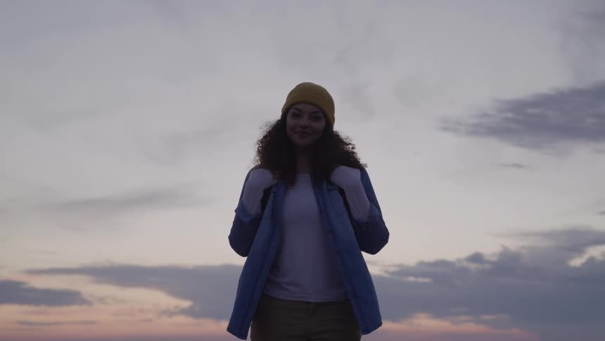 A young woman stands outdoors wearing a jacket and holding the straps of her backpack wearing a hat and smiling under a cloudy sky at sunset