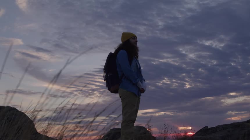 A woman stands in a field carrying a backpack holding her own hands in front of her looking to the ground at first then looking up to the sky