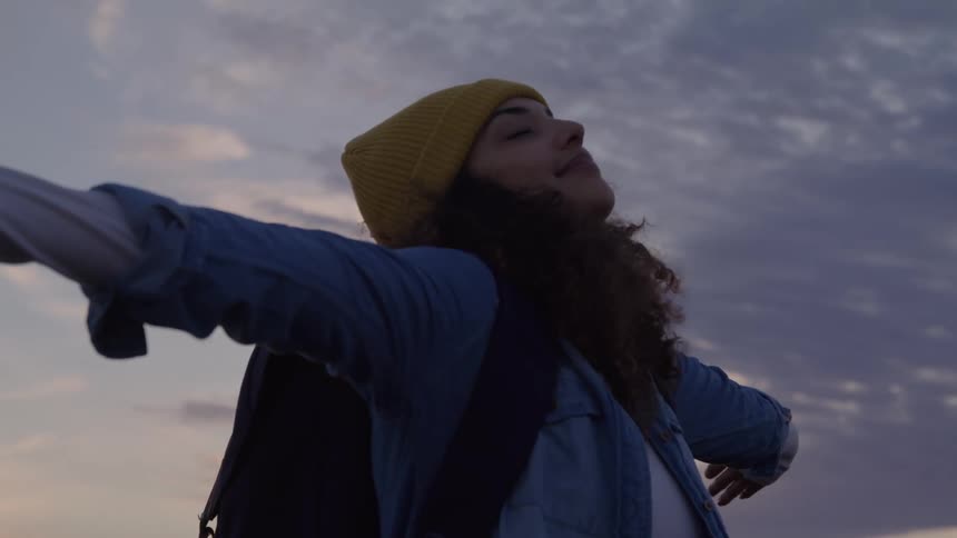 A joyful woman wearing a yellow beanie and a backpack stands on a mountain ridge and raises her arms up with her head tilted back and her eyes closed as she looks up at a cloudy sunset sky