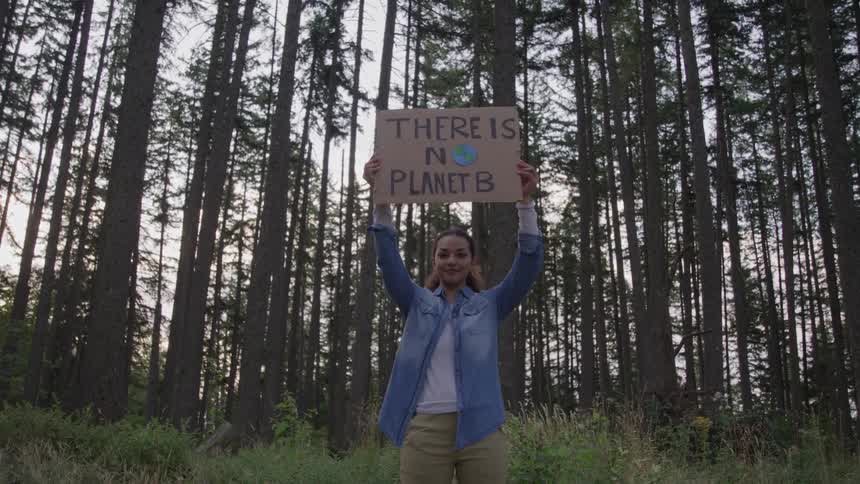 A woman standing in a forest clearing smiling as she holds up a sign saying there is no planet b