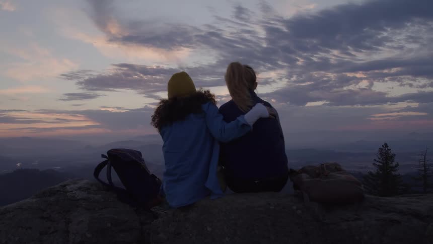 Two women sitting on a mountain peak embracing each other with a sunset view