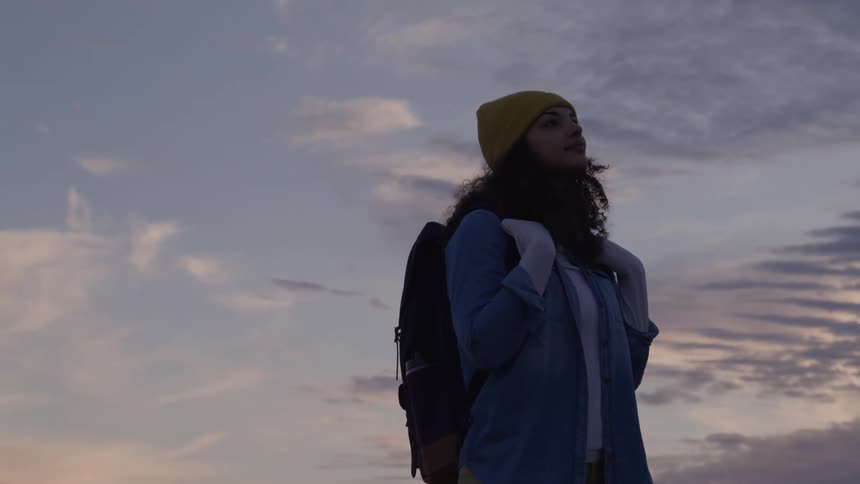 A young female hiker wearing a yellow beanie a blue denim jacket and a black backpack is standing on a mountain ridge and looking out at the sunlit cloudy sky