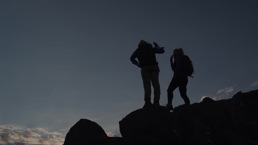 A male hiker is climbing up a rocky mountain ridge toward his female hiking partner who reaches down to help him up and then they stand together and raise their hands in a celebratory high five