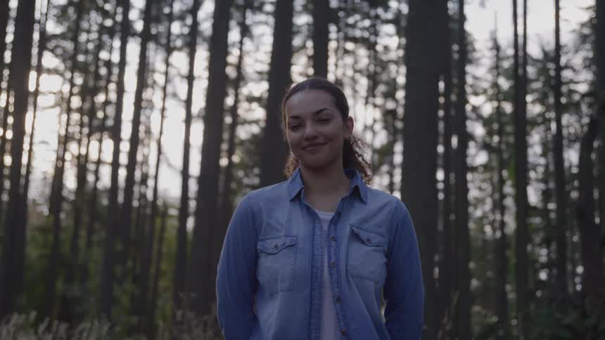 A happy woman wearing a light blue denim shirt over a white tshirt and with her curly hair in a ponytail is standing in a forest smiling and looking at the viewer as the sun shines through the tall trees behind her