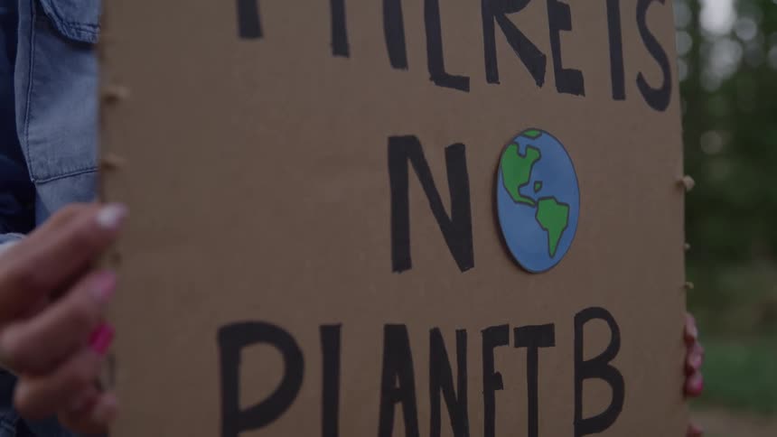 A protestor wearing a denim shirt is holding a cardboard sign that reads there is no planet b with a picture of the earth on it while standing in a blurry green forest