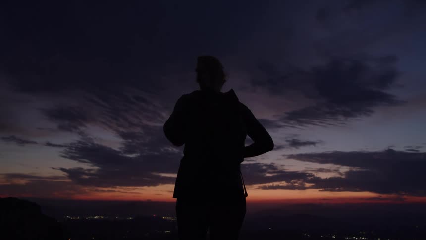 A silhouetted person wearing a backpack stands on a mountain ridge with their hands on their hips looking out over a city with distant lights as the sky turns a dark mix of blue purple and orange