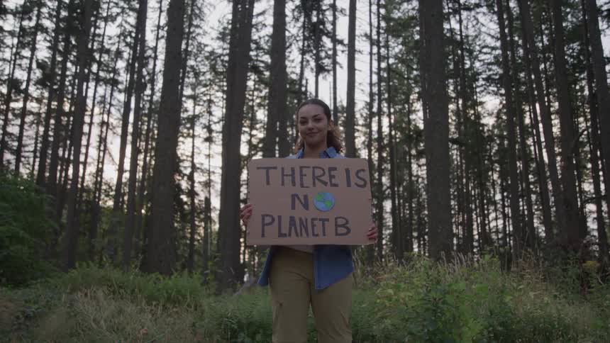 A young woman in the woods holds a placard that says there is no planet b surrounded by trees at midday