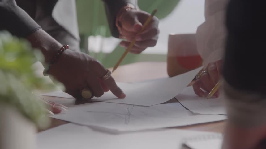The hands of several professionals are gathered around a table and pointing at papers with technical drawings with one person holding a pencil and another wearing several rings and bracelets