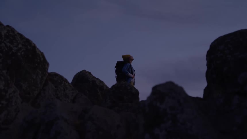 A hiker wearing a yellow beanie and a backpack stands on a rocky outcrop looking out at a valley below with the deep blue sky and dark clouds in the background