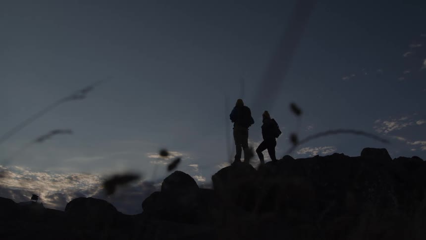 Two women standing on top of a rock hiking through a rocky terrain in the evening both carrying backpacks