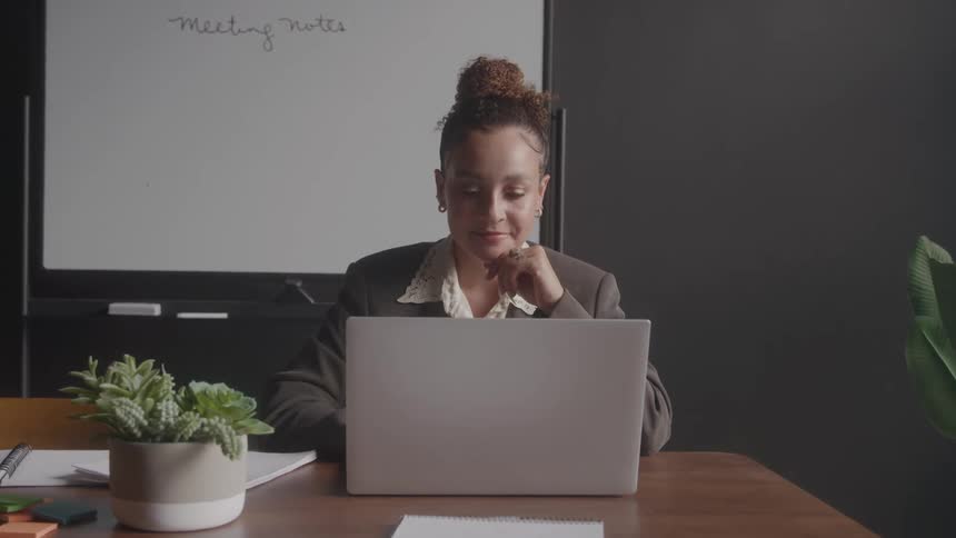 A professional woman with her curly hair in a bun is sitting at a desk and looking at her laptop screen with her chin resting on her hand with a plant and notebooks on the desk and a whiteboard in the background