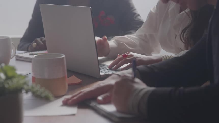 Three people sit at a coffee table in an office being taught by the woman in the middle who is typing on a laptop and gesturing with her right hand while the other two men are busy writing at midday
