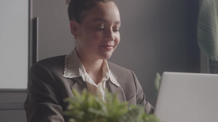 A professional woman with her curly hair tied in a bun is smiling while she looks at her laptop screen with a green potted plant blurred in the foreground