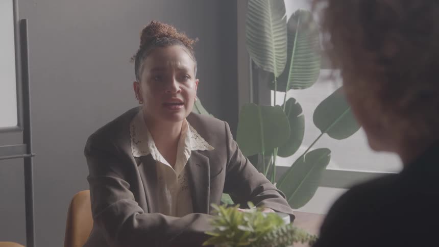 A young woman wearing a blazer conducts an interview with a man inside an office at midday