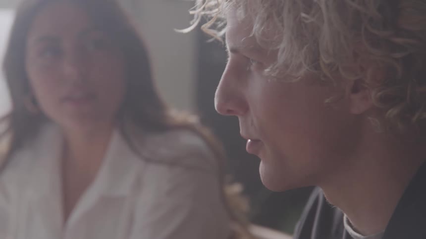 A young man with curly blonde hair and is seen in a profile view talking next a blurred woman in the background