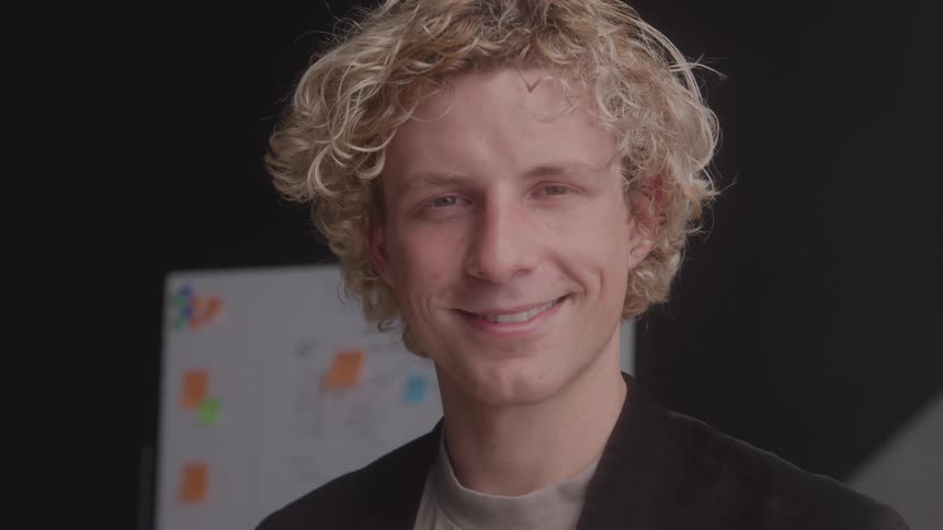 Blond curlyhaired man smiling with a blurred whiteboard in the background