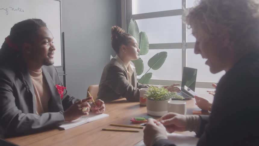 Four people inside a conference room are all discussing with a woman sitting and typing on her laptop while the others are busy with other activities at midday