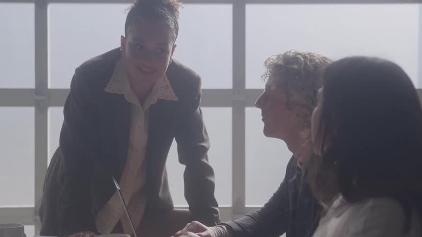 Woman standing and talking to two seated colleagues in a meeting lit by natural light