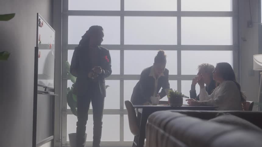 A team of four coworkers is in a brightly lit office with one standing and writing on a whiteboard while the others three sit at a table