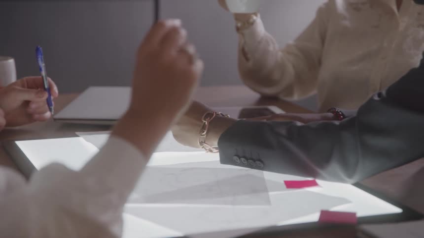 Three people sitting by a table with papers over a light box near a tablet and a closed laptop discuss a project and take notes while one of them holds up a mug