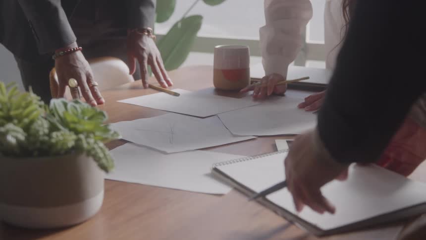 The hands of several professionals are gathered around a table with papers with one person writing with a pencil and another pointing with a pen as a plant and mugs sit nearby