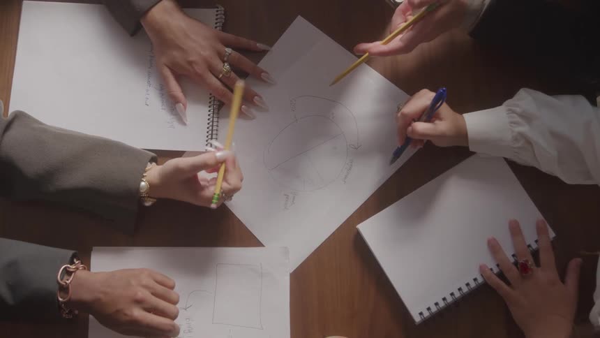 A topdown view shows several peoples hands some wearing rings and bracelets working together with pencils and a pen to draw diagrams on sheets of paper on a wooden table