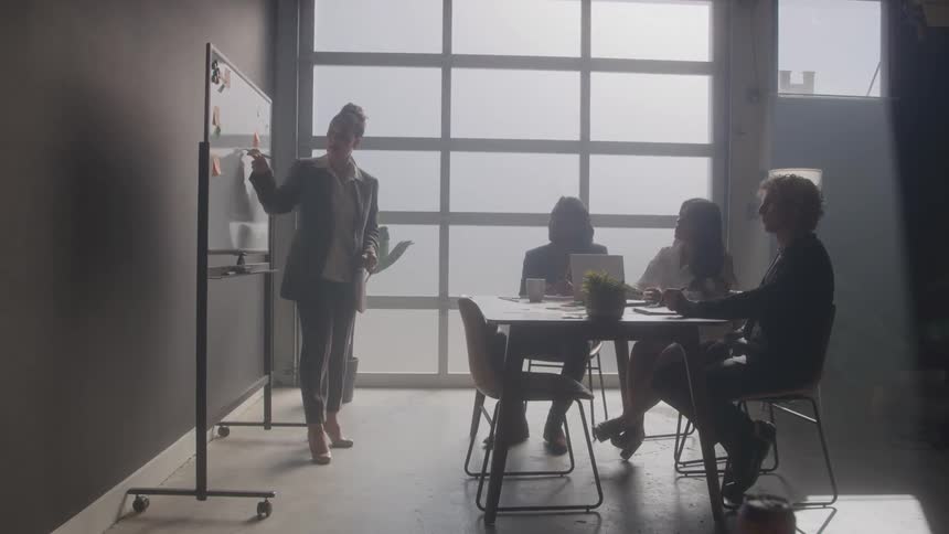 A female lecturer teaching three students seated behind a coffee table with a whiteboard inside a small space at midday