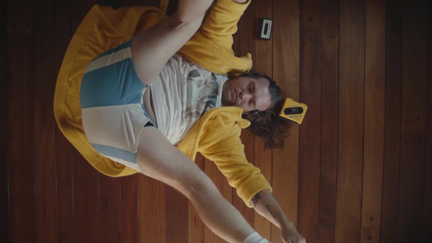 A man with dark hair is lying on his back on a wooden floor wearing a yellow robe and a white shirt with a graphic on it stretching his legs with his arms while a walkman and a cassette are on the floor near him