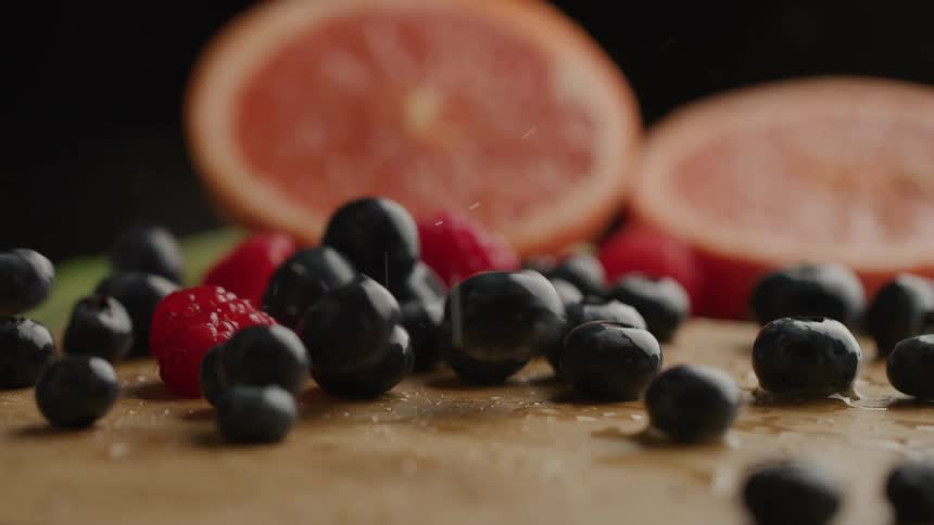The video shows a closeup of raspberries and blueberries on a wooden surface with grapefruit halves in the background several blueberries bounce and roll onto the surface joining the other fruit