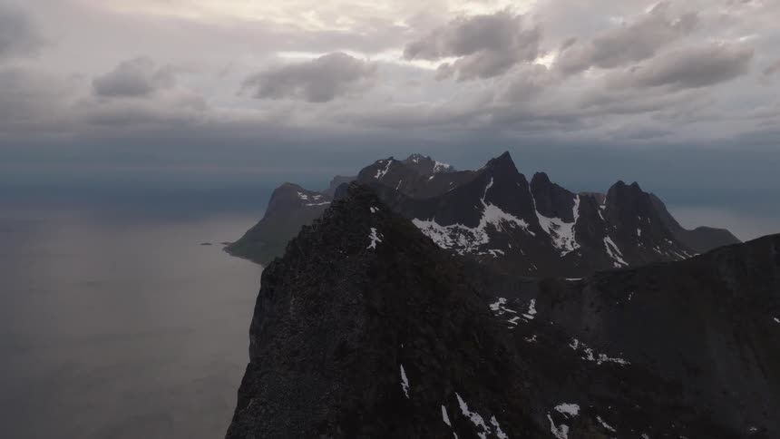 A dramatic mountain range with sharp jagged peaks and patches of snow is shown from above with dark clouds in the sky and a large body of water on the left side of the frame