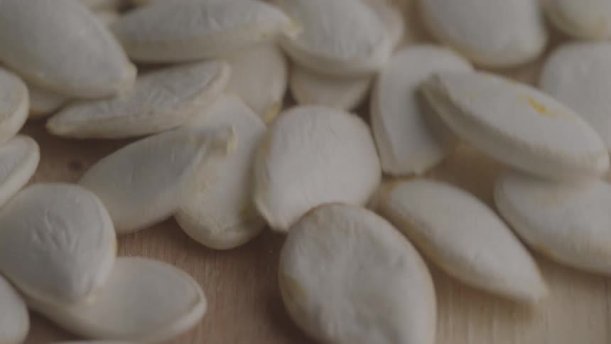 A closeup view shows several white pumpkin seeds falling from a wood surface