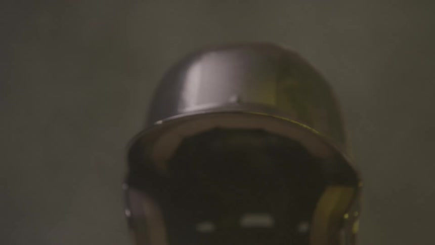 A dark glossy baseball helmet slowly rotates on a stand illuminated by shifting purple and yellow lights in a studiolike setting