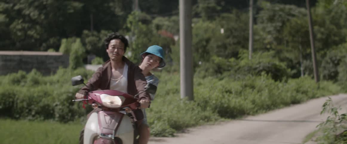 A man and a woman riding a motorcycle down a dirt road and her hat falls off