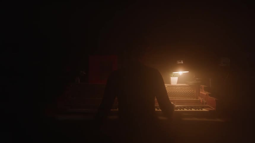 The video shows a dark room with a single lamp illuminating a vintage organ a persons silhouette is visible standing in the foreground with their back to the camera
