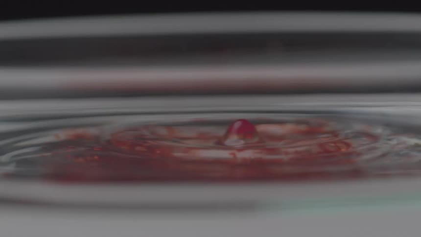 A close view of a clear liquid shows drops of red liquid falling into it and staining the water as ripples slowly spread across the surface