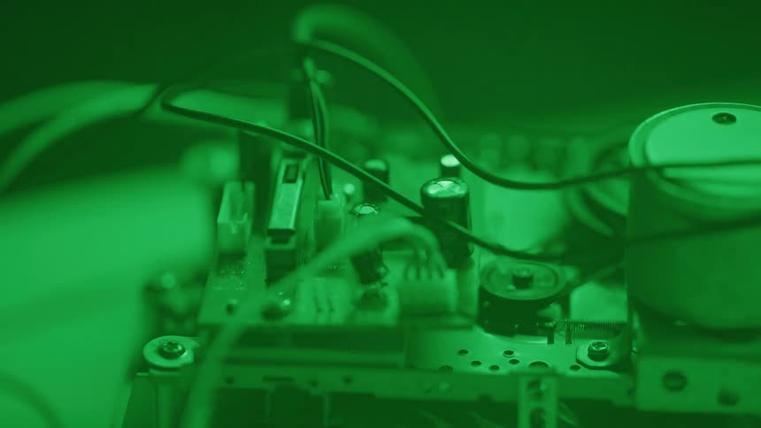 A close view shows the inner electronic components of a machine with various wires and a circuit board all illuminated by a soft green light