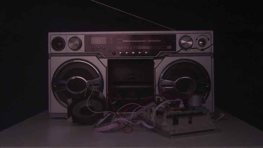 A view of an old silver boom box shows it sitting on a white table with components exposed on the right side and a speaker detached on the left side as the lights on the front of the radio pulse with a beat