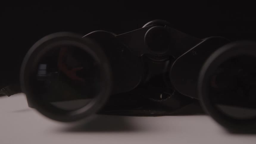 A pair of black binoculars rests on a light surface with reflections shimmering in their lenses