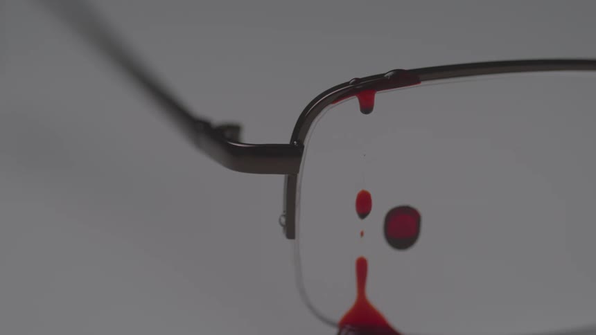 Red liquid drips and smears across the lens and frame of a pair of glasses against a plain background