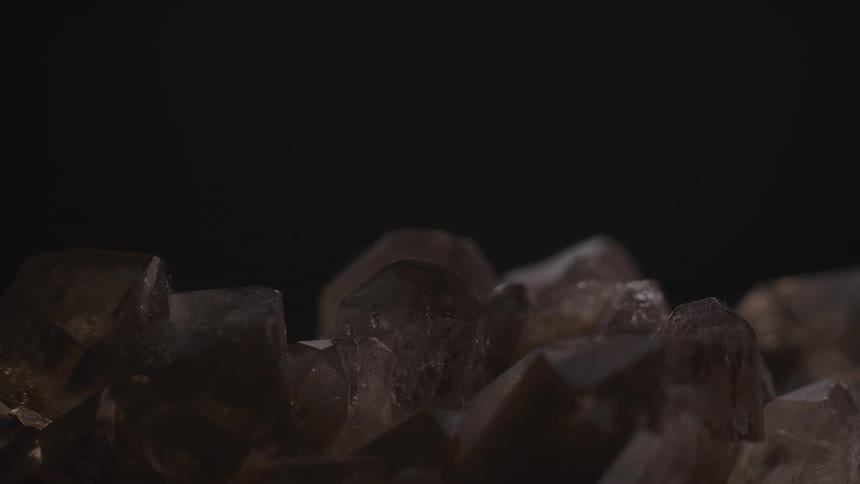 A collection of dark translucent crystals with pointed facets is gradually illuminated from the top revealing their shimmering surfaces